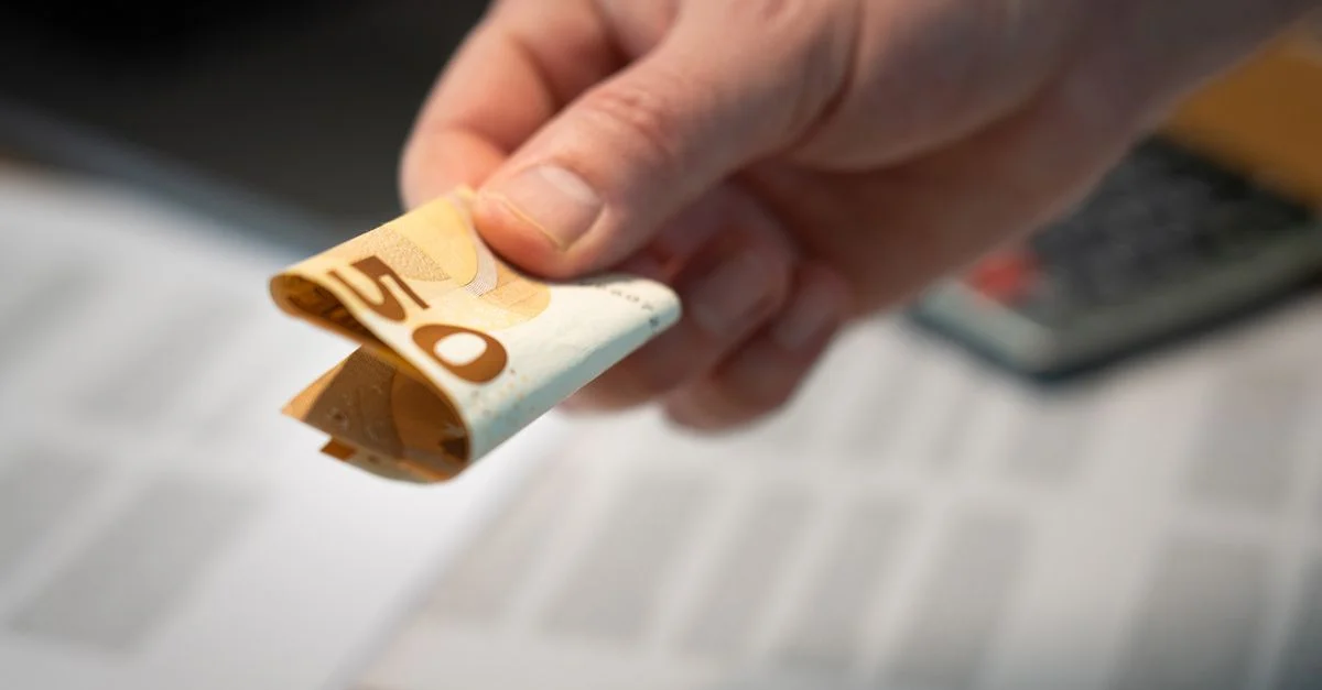 Une image détaillée d'une main tenant un billet de 50 euros plié, avec des documents en arrière-plan.